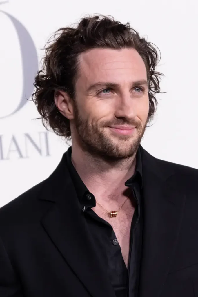 Person in a black suit on a red carpet, with neatly styled wavy hair and a subtle smile
