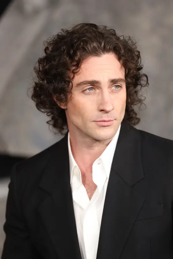 Person wearing a formal suit with curly hair, looking to the side at an event