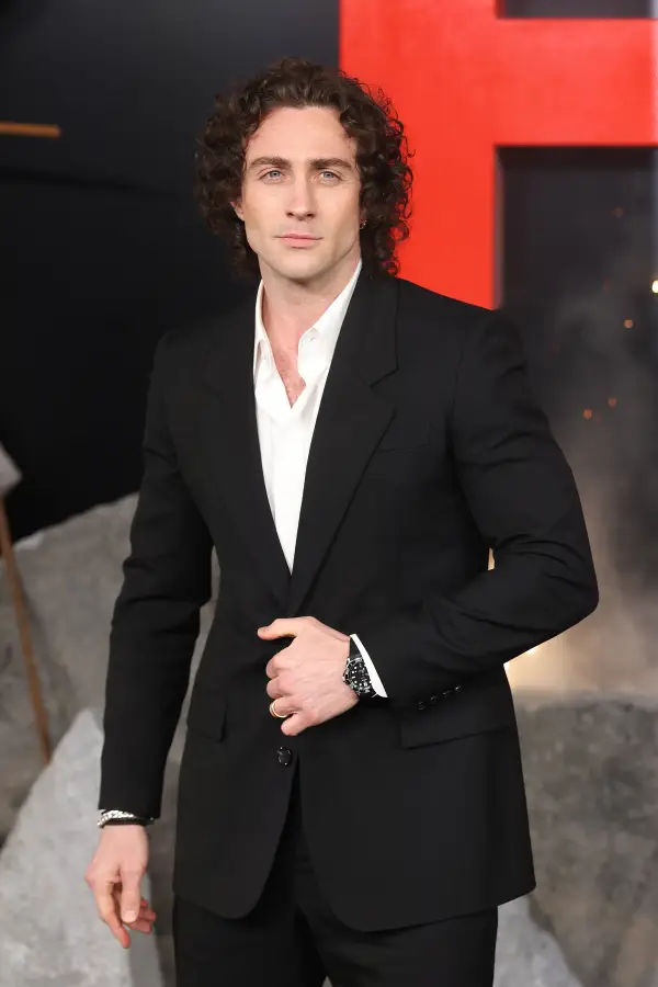 Person at a formal event wearing a black suit and white shirt, posing with one hand adjusting the jacket