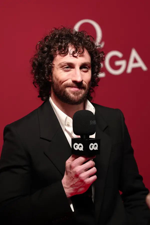 Person in a black suit holding a GQ microphone on a red carpet with a background logo