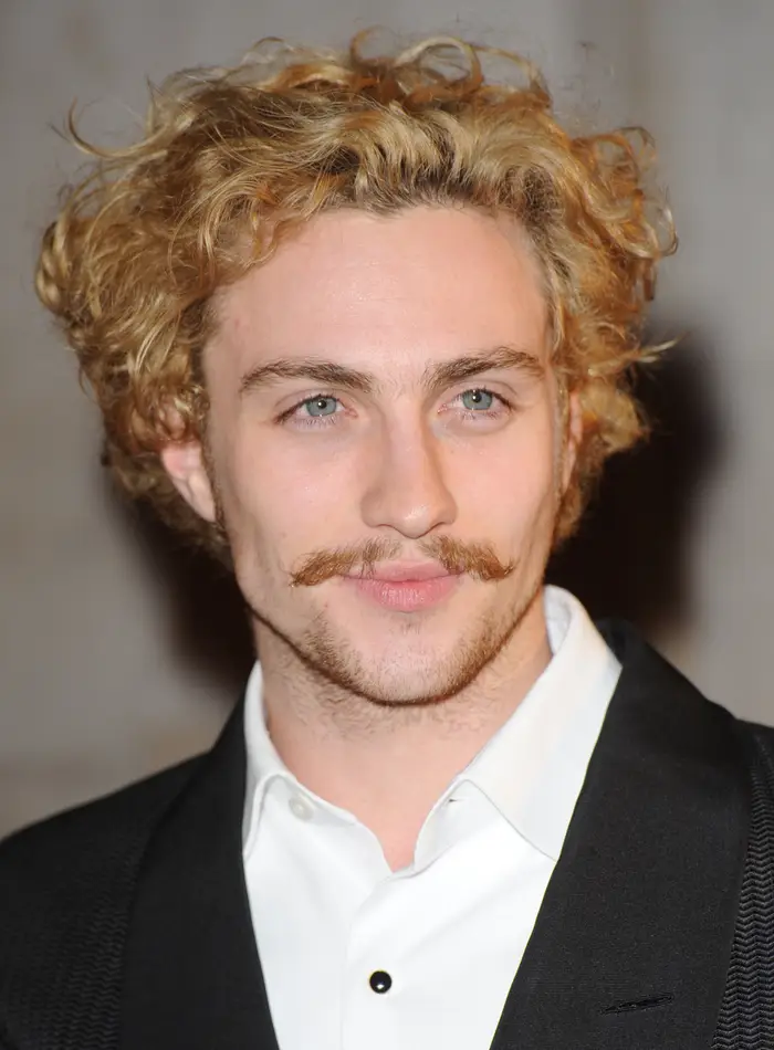 Person with curly hair and mustache, wearing a suit with a white shirt, smiles slightly, looking away from the camera
